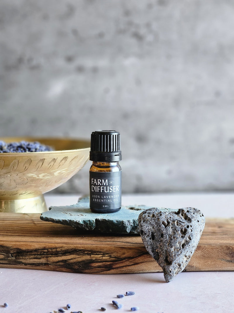 Lavender Essential Oil sitting on a green volcanic rock, sitting on a piece of olive wood, with a heart shaped lave rock in the foreground and a bowl of lavender buds in the background.