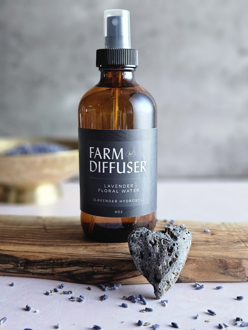 Lavender Floral Water, 100% Lavender Hydrosol on an olive wood slab, with a heart shaped lava rock and lavender buds in the foreground, with a brass bowl of lavender buds ion the background